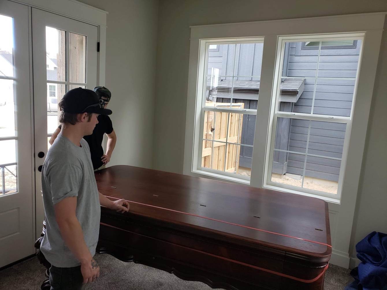 Two Delicate Touch movers lowering a long dark polished wood dining table into place in an empty, freshly painted room of a new build, a red lifting strap still running the length of the tabletop and a blue moving blanket on the carpet beside it