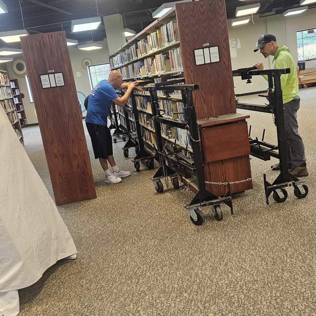 Two Delicate Touch movers rolling fully loaded library shelving on steel book-cart dollies, with the wooden end panels unbolted and set aside