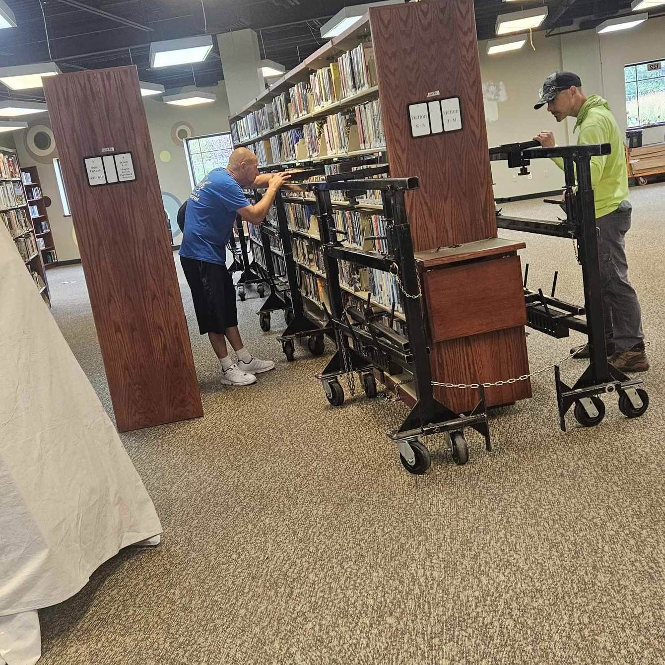 Two Delicate Touch movers rolling fully loaded library shelving on steel book-cart dollies, with the wooden end panels unbolted and set aside