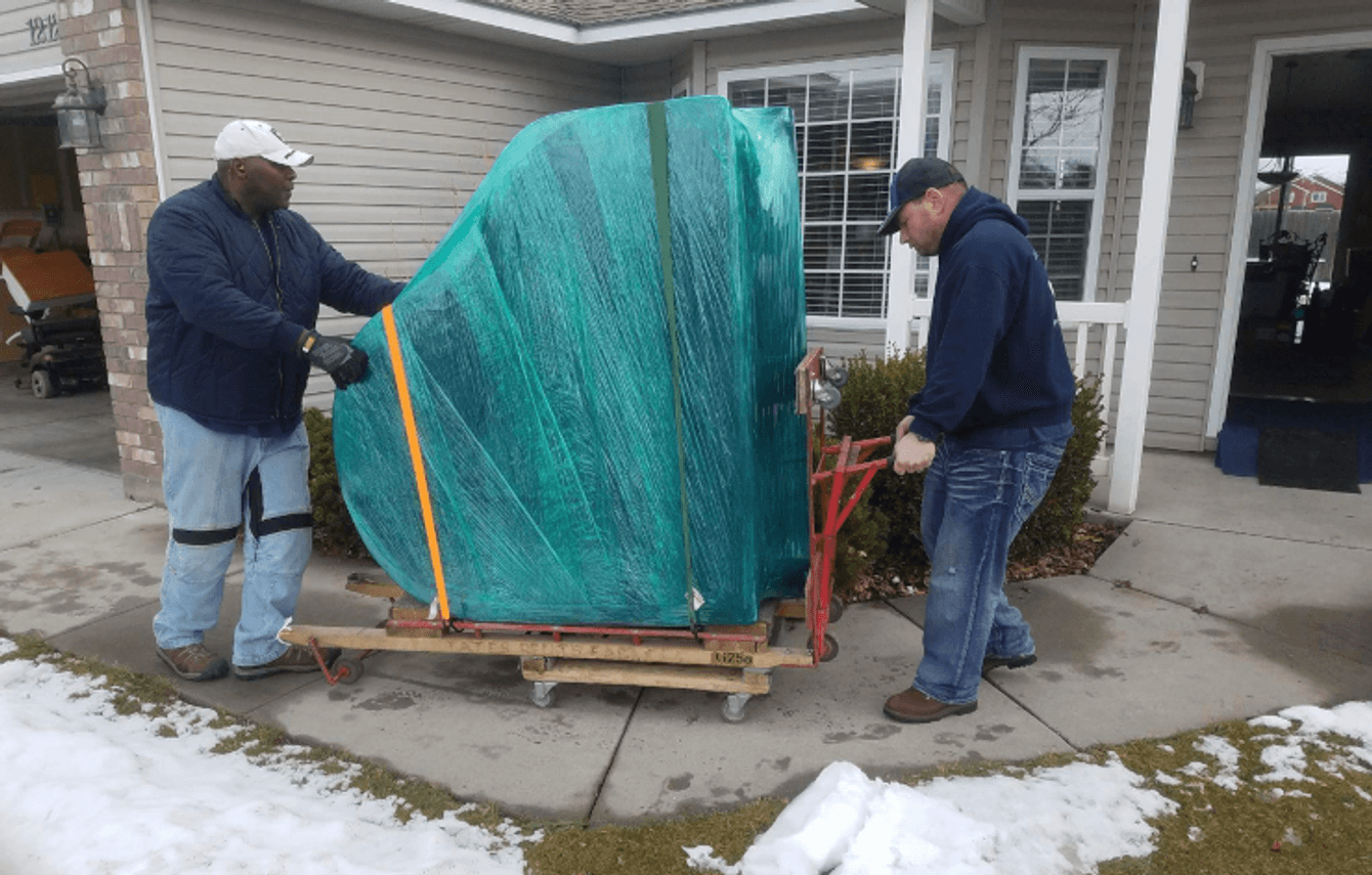 Two Delicate Touch movers steadying a piano wrapped in green stretch film and yellow straps on a wheeled wooden board, on a house walkway with snow still on the lawn