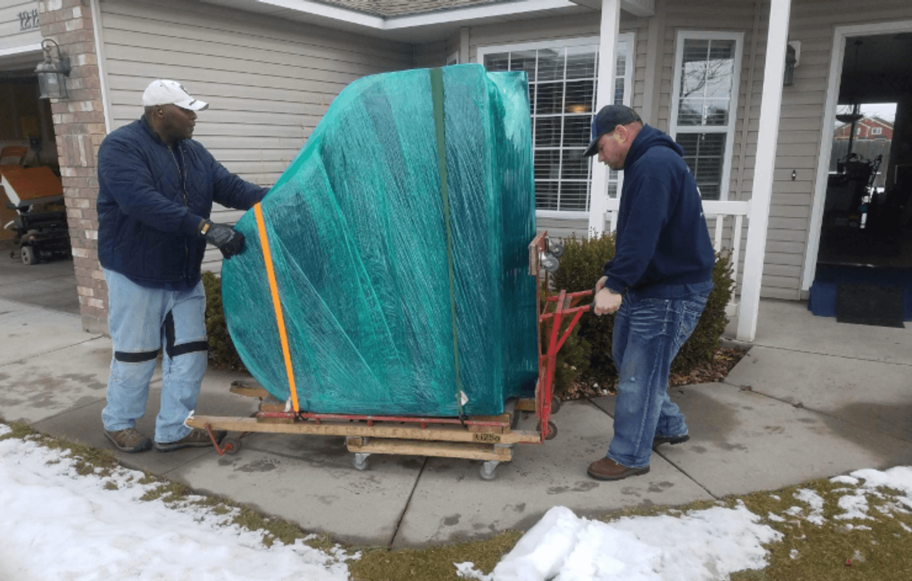 Two Delicate Touch movers steadying a piano wrapped in green stretch film and yellow straps on a wheeled wooden board, on a house walkway with snow still on the lawn